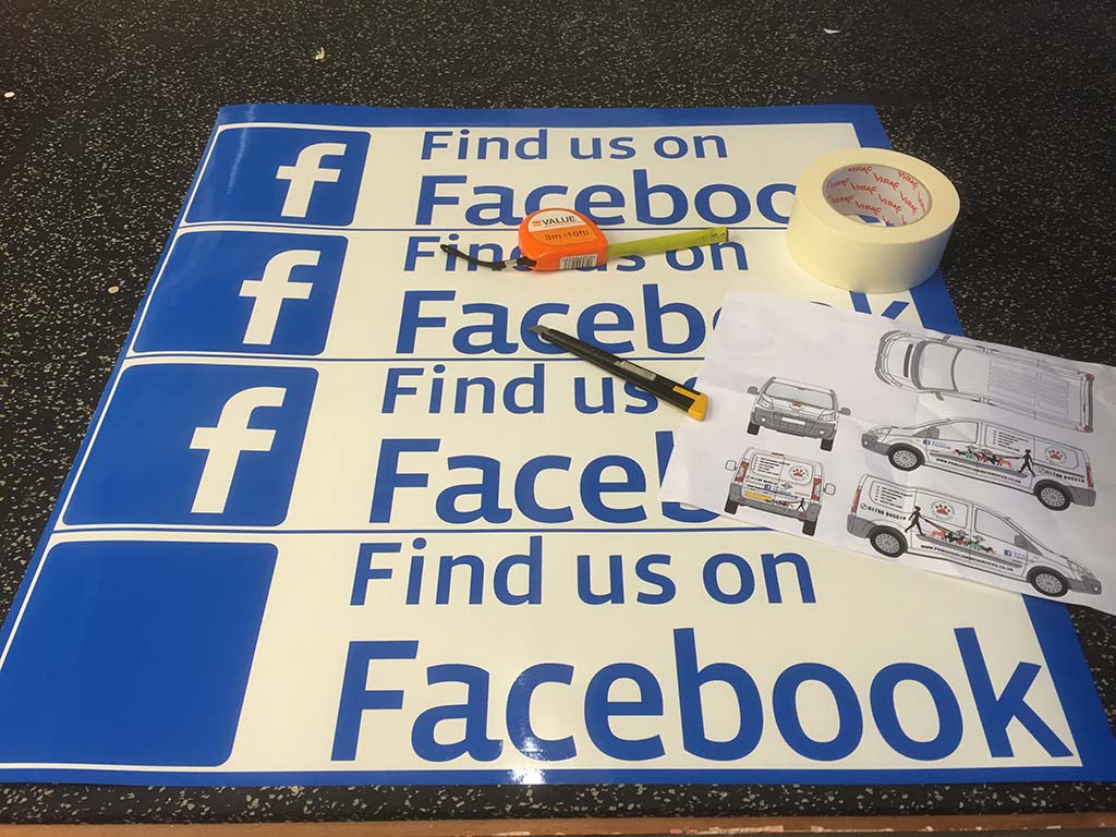 sheet of cut and partialy weeded blue vinyl facebook graphics lying on desk with measuring tape, masking tape, olfa graphic knife, and van layout drawing
